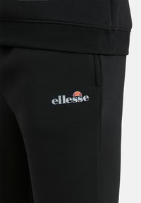 Zwarte sweatpants met een elastische tailleband, zijzakken en een bedrukte witte en rode "ellesse" logo op de dij. Gladde textuur.