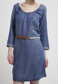 Nainen, joka on pukeutunut polvipituiseen, siniseen farkkuvermeeseen, jossa on 3/4-sleeves, rintatasku ja kaksivärinen ruskea ja valkoinen punottu vyö.