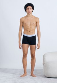 Mustad boxeri-stiilis aluspüksid heledat halli vöökohaga, millel on logo tekst. Modell poseerib jalul heleda vaibakattega pinnal.