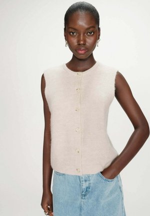 Femme à la peau foncée portant un haut sans manches beige boutonné et un jean denim bleu clair, debout avec une main dans la poche.