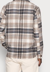 Man in een beige, grijs en zwart geruit overhemd met lange mouwen, met crèmekleurige broek, gezien van achteren tegen een eenvoudige achtergrond.