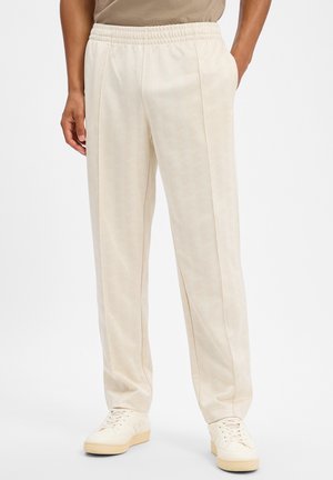 Mann trägt beige elastische Bundhose und passende cremefarbene Sneaker, steht mit teilweise in den Taschen steckenden Händen.