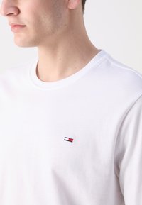 Camiseta blanca de algodón con cuello redondo, que presenta un pequeño logotipo bordado en rojo, azul y blanco en la parte izquierda del pecho. Textura suave.