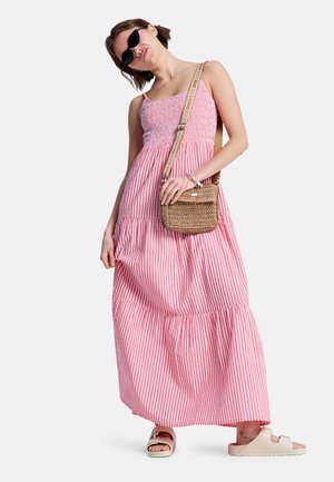 Kobieta w różowo-białej sukience maxi w paski, czarne okulary przeciwsłoneczne, nosząca plecioną torbę na ramię i beżowe sandały, stojąca na białym tle.