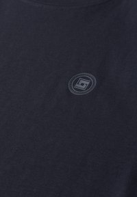 Tissu bleu marine foncé avec un petit logo circulaire présentant des lettres stylisées centré près de l'encolure.