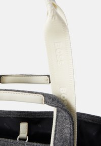 Sivi filcasti kovčeg s krem kožnim detaljima i ručkama. Ima teksturirani remen s logotipom "BOSS". Unutrašnjost izgleda tamno i podstavljena.