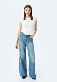Witte top met korte mouwen en naaddetails, gecombineerd met lichtblauwe wijde jeans. Model draagt zwarte puntige hakken op een effen achtergrond.