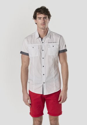 Fehér rövid ujjú, gombos ing két mellzsebbel, sötétkék díszítéssel és fekete gombokkal, piros rövidnadrággal párosítva.