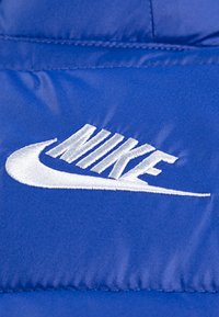 Mėlynas audinys su išsiskiriančiu balto siuvinėjimo Nike logotipu, kuriame matomas kreivai išlenktas swoosh, demonstruojantis lygią tekstūrą ir tolygų siuvimą.