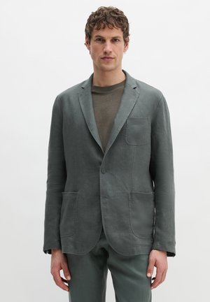 Mann trägt ein graues Sakko mit einem Knopf über einem dunkelgrauen Rundhalsausschnittshirt, steht vor einem schlichten weißen Hintergrund.