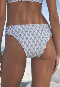 Weißes Bikini-Höschen mit blauem Dekorationsmuster, gesäumt mit Rüschen. Glatte Textur, mittelhoher Schnitt, geeignet für Strandmode.
