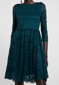 Teal Spitzenkleid mit langen transparenten Ärmeln, tailliertem Oberteil und schwingendem Rock. Blumiges Spitzenmuster und wellenförmige Saumverzierungen.