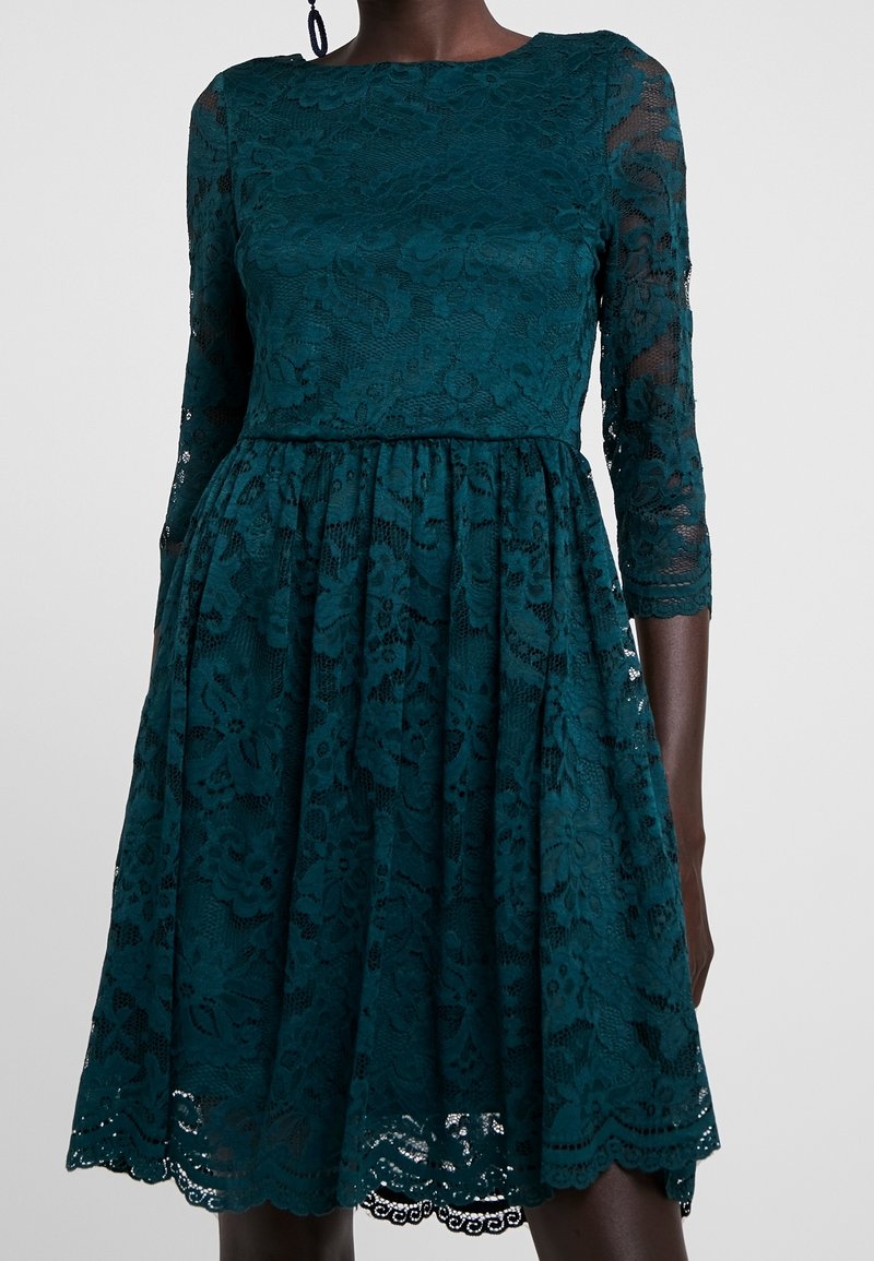 Teal Spitzenkleid mit langen transparenten Ärmeln, tailliertem Oberteil und schwingendem Rock. Blumiges Spitzenmuster und wellenförmige Saumverzierungen.