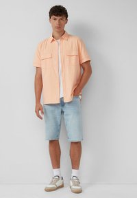 Korte mouwen, licht perzik kleurige button-up shirt met twee voortassen, gecombineerd met lichtblauwe denim korte broek en beige sneakers.