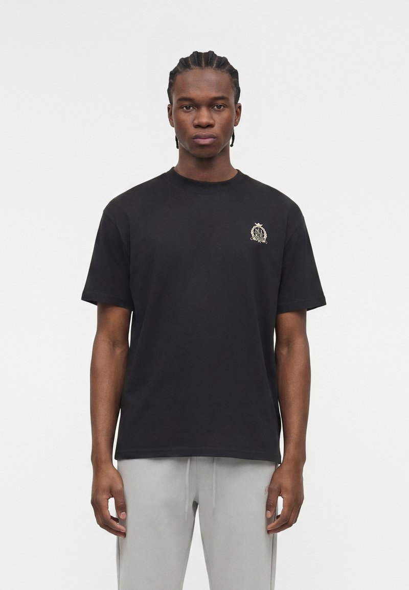 Giovane uomo con capelli intrecciati che indossa una maglietta nera con un piccolo logo sul petto e pantaloni grigio chiaro, in piedi davanti a uno sfondo semplice.