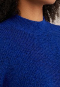 Maglione lavorato a maglia blu con un collo alto, caratterizzato da una superficie testurizzata e un aspetto morbido e peloso. Nessun motivo o hardware visibile.