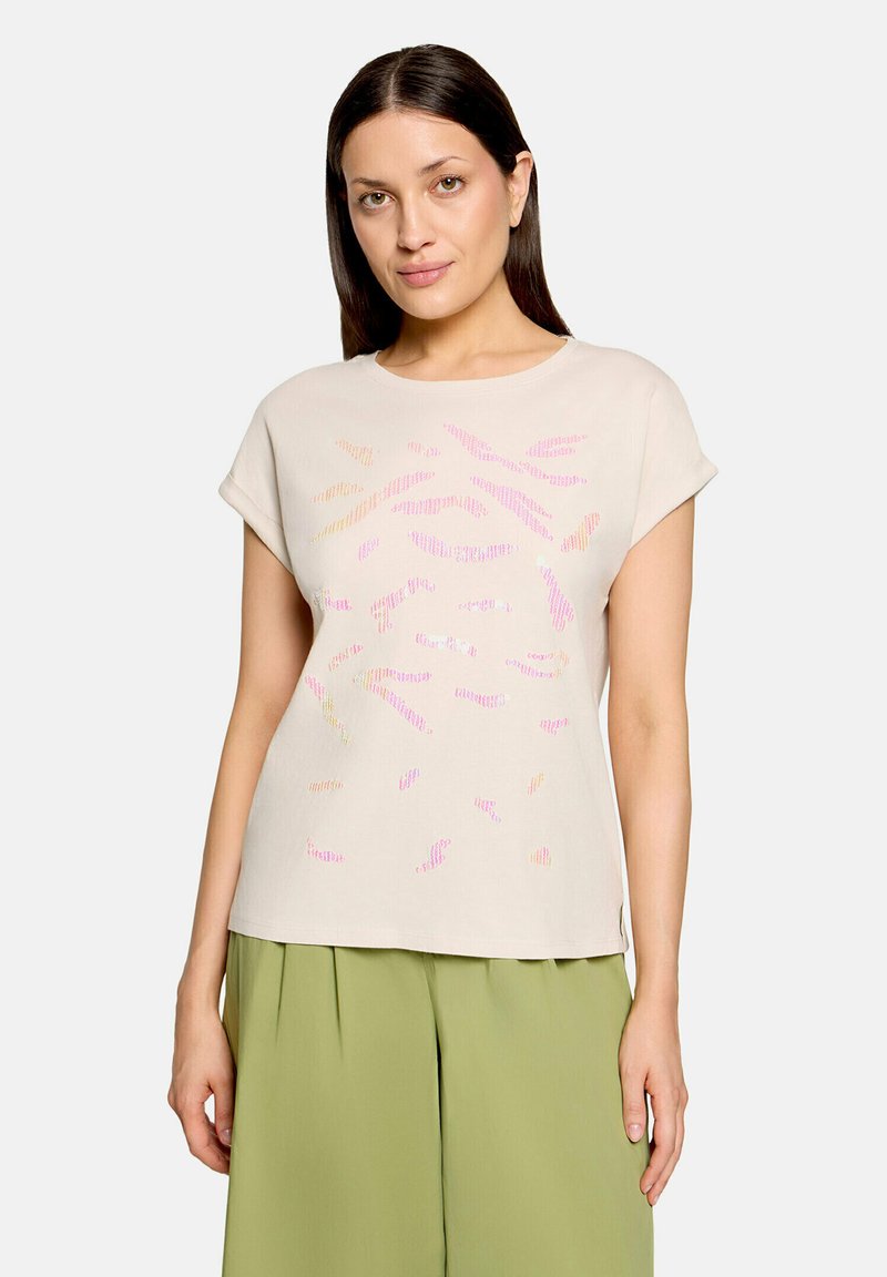 Femme aux cheveux raides et foncés portant un haut beige à manches courtes avec des imprimés abstraits roses et orange et un pantalon ample vert clair.