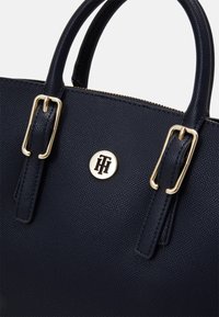 Borsa a tracolla in pelle texturizzata blu navy con doppie maniglie, fibbie dorate, chiusura con cerniera e un emblematico logo dorato rotondo sul davanti.