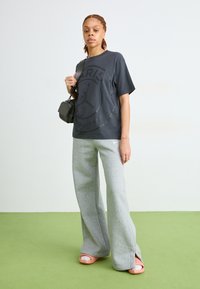 Grijze oversized T-shirt met een zwart logo, gecombineerd met grijze wijde sweatpants en roze slippertjes. Gecompleteerd met een kleine zwarte tas.