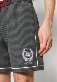 Gros plan de la main d'une personne à côté d'un short gris à fines rayures avec un liseré blanc et un patch logo "Hugo est. 1993" sur la jambe gauche.