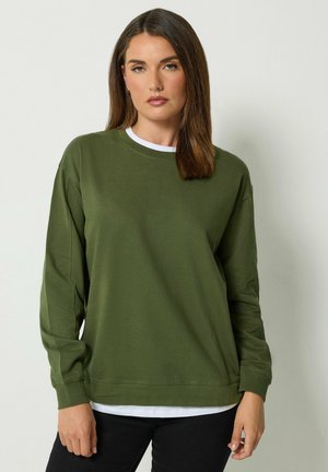 Donna con capelli castani lisci che indossa una camicia verde oliva a maniche lunghe sopra una maglietta bianca e pantaloni neri, in piedi davanti a uno sfondo chiaro e semplice.