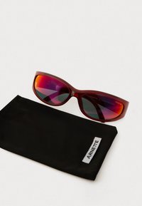 Crvene Arnette naočale sa reflektirajućim ljubičasto-narančastim lećama pored crnog mekanog futrola označenog s Arnette na bijeloj površini.