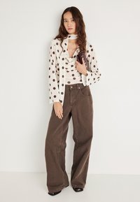 Bluzka z długim rękawem w brązowe kropki, zestawiona z brązowymi szerokimi spodniami. Dodatkiem jest mała bordowa torebka. Jednolite tło.
