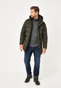 Dunkelgrüne Pufferjacke mit Kapuze, grauer Rollkragenpullover, blaue Jeans und schwarze Stiefel. Verfügt über gerippte Bündchen und dezente Karomusterakzente.