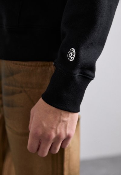 Sweatshirt noir à poignets côtelés, arborant un petit patch logo sur la manche gauche. Pantalon marron avec une surface texturée visible en arrière-plan.