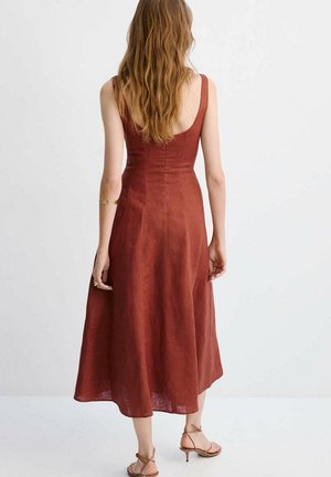 Mujer con cabello largo vestida con un vestido midi sin mangas de color óxido y sandalias de tacón con tiras, de pie frente a un fondo liso y claro.