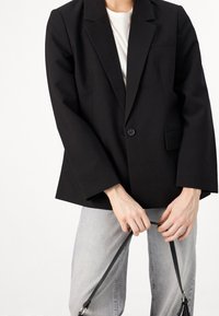 Personne portant un blazer noir sur une chemise blanche et un jean gris clair, tenant avec les deux mains les bandes noires du sac devant elle.