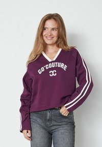 Jauna sieviete ar garām matēm, ģērbusies purpura krāsas džemperī ar baltām svītrām uz piedurknēm un tekstu "CO'COUTURE", smaidot pozē ar roku kabatā.