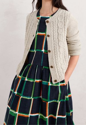 Kobieta w kremowym kardiganie na guziki o wzorze splotu warkoczowego, nałożonym na granatową sukienkę z zielonymi, pomarańczowymi i białymi kwadratowymi wzorami, z rękami w kieszeniach.