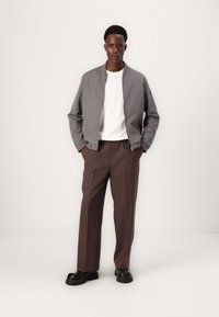 Gray bomber jacket with a zip front, white T-shirt, and brown textured pants. Black chunky-soled shoes complete the outfit.