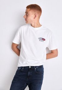 Balta kokvilnas T-kreklā, kas rotāts ar sarkanu un zilu logotipu ar tekstu "TOMMY Worldwide". Apvienots ar tumšu denima džinsu.