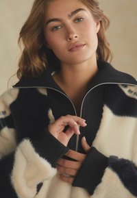 Fliisist zip-jakk, millel on mustad ja kreemjad abstraktsed mustrid, kõrge krae, soonilised varrukad ja sile, pehme tekstuur.