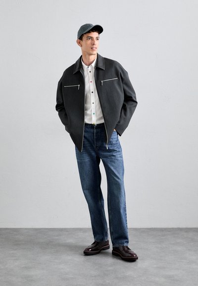 Grå ulljakke med to fremre glidelåslommer, hvit skjorte med flerfargede knapper, blå jeans og mørkebrune sko.