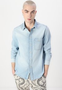 Lys blå denimskjorte med spredt krage, lange ermer, brystlomme og synlig knappeplacket, kombinert med mønstrete beige bukser.