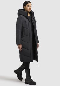 Sort pufferjakke med hætte, prydet med et langt design, chevron-sting og sidelommer. Bruges med sorte combat-støvler.