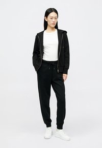 Čierna mikina na zips so bočnými vreckami, nosená cez biely tričko, doplnená čiernymi teplákmi. Biely tenisky dotvárajú vzhľad.