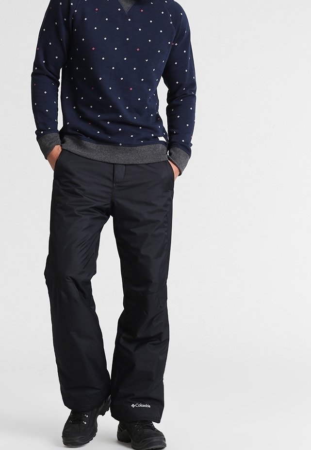 Homme portant un pantalon de neige Columbia noir, des chaussures noires et un pull bleu marine avec de petits motifs losanges blancs et rouges, mains dans les poches.