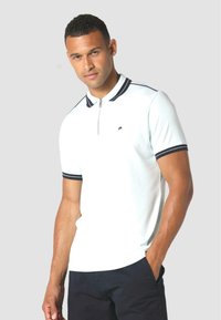 Baltas polo krekls ar melniem akcentiem, īsām piedurknēm un rāvējslēdzēja aizdari. Mazs logo kreisajā krūtī. Nēsāts ar tumšām biksēm.