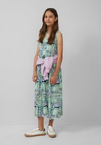 Buntes knielanges Kleid mit abstrakten geometrischen Mustern in Grün, Lila und Schwarz. Kombiniert mit weißen Sneakern und einem rosa Pullover, der um die Taille gebunden ist.