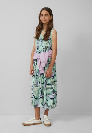 Buntes knielanges Kleid mit abstrakten geometrischen Mustern in Grün, Lila und Schwarz. Kombiniert mit weißen Sneakern und einem rosa Pullover, der um die Taille gebunden ist.