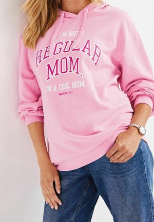 Vrouw draagt een roze hoodie met de tekst "I'm not a regular mom. I'm a cool mom. Mean Girls," gecombineerd met een blauwe spijkerbroek en een zilveren horloge.