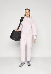 Rosa Sweatshirt mit Logo, passende Jogginghosen, graue Turnschuhe und eine schwarze Sporttasche. Das Outfit ist lässig und bequem.