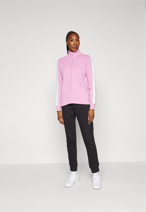 Giacca rosa con zip e maniche bianche, dotata di tasche e logo, abbinata a pantaloni neri e scarpe da ginnastica bianche. Trama del tessuto liscia.