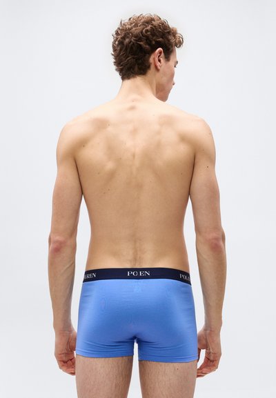 Άνδρας μοντέλο που στέκεται γυρισμένος ανάποδα, φοράει μπλε μποξεράκια με σκούρα μέση και με το λογότυπο "Polo Ralph Lauren".