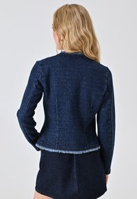 Giacca in denim blu scuro con finitura testurizzata, caratterizzata da maniche lunghe e dettagli in frange lungo l'orlo. Vista posteriore mostrata.