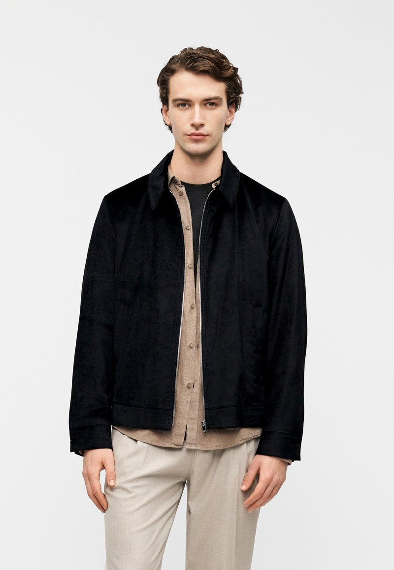 Giacca bomber in pelle scamosciata nera con zip frontale e colletto, indossata sopra una camicia marrone chiaro con bottoni, abbinata a pantaloni beige.